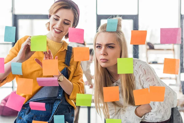 Beautiful casual businesswomen looking at colorful sticky notes on glass window in office — Stock Photo