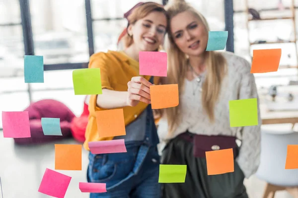 Beautiful casual businesswomen colorful putting sticky notes on glass window in office — Stock Photo