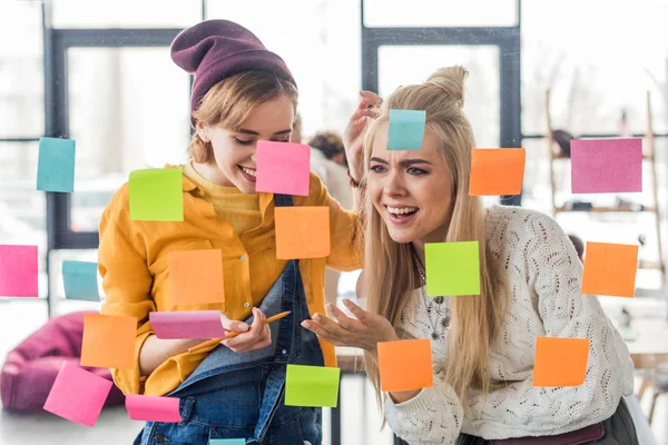Schöne Gelegenheitsunternehmerinnen lachen neben bunten klebrigen Zetteln an Glasfenstern im Büro — Stockfoto