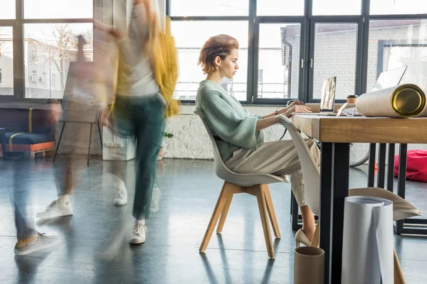 Female architect sitting and using laptop at desk in loft office with colleagues in motion blur on background — Stock Photo
