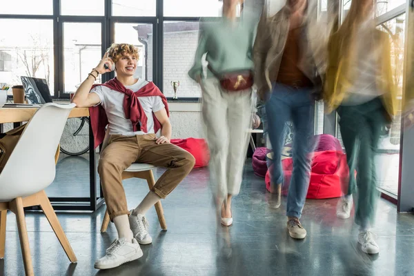 Sorridente empresário casual em fones de ouvido sentado no escritório loft com colegas em movimento borrão no fundo — Fotografia de Stock