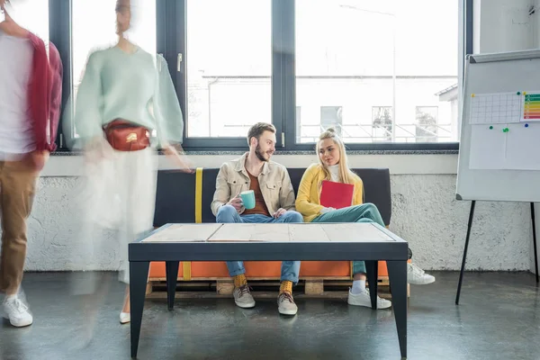 Female and male casual businesspeople sitting on couch and having discussion in loft office with colleagues in motion blur on background — Stock Photo