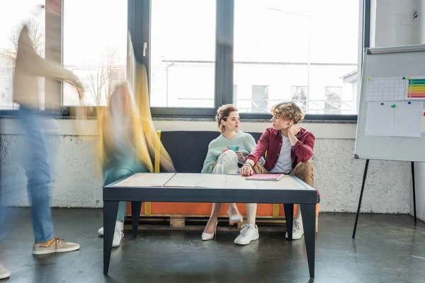 Hombres y mujeres empresarios ocasionales sentados y teniendo discusión en la oficina loft con colegas en movimiento desenfoque en el fondo - foto de stock