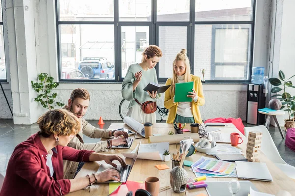 Grupo enfocado de diseñadores femeninos y masculinos que trabajan en el proyecto de startup con computadoras portátiles y planos en la oficina loft - foto de stock