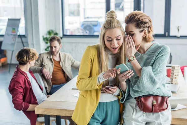 Mulheres de negócios casuais bonitas sussurrando e usando smartphones no escritório loft com colegas em segundo plano — Fotografia de Stock