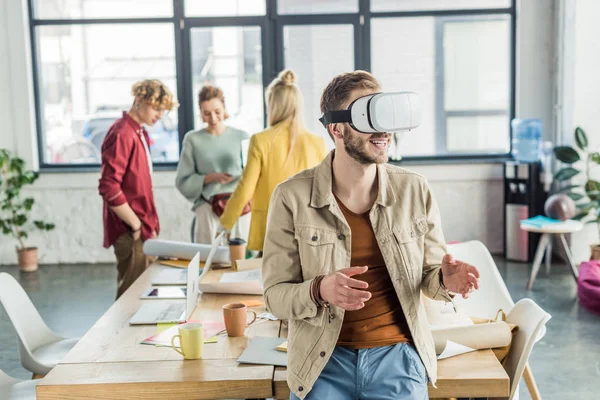 Sonriente diseñador masculino gesto con las manos mientras tiene experiencia de realidad virtual en la oficina loft con colegas de fondo - foto de stock