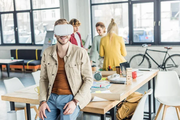 Diseñador masculino sonriente que tiene experiencia de realidad virtual en la oficina loft con colegas de fondo - foto de stock