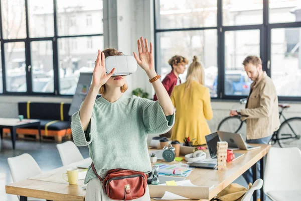 Female designer gesturing with hands while having virtual reality experience in loft office with colleagues on background — Stock Photo