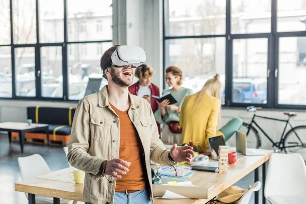 Aufgeregter männlicher Designer gestikuliert mit den Händen, während er Virtual-Reality-Erfahrung im Loft-Büro mit Kollegen im Hintergrund hat — Stockfoto