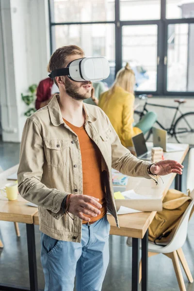Männlicher Designer gestikuliert mit den Händen, während er Virtual-Reality-Erfahrung im Loft-Büro mit Kollegen im Hintergrund hat — Stockfoto