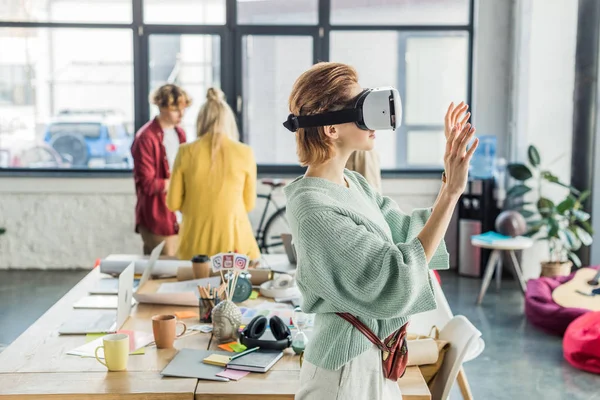 Designer feminino gesticular com as mãos enquanto tendo experiência realidade virtual no escritório loft com colegas em segundo plano — Fotografia de Stock