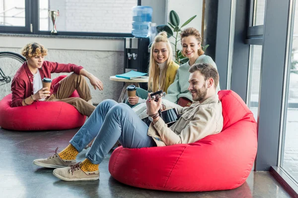 Groupe souriant d'amis assis sur des chaises de sac de haricot et jouer de la guitare — Photo de stock