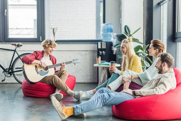 Grupo de amigos sentados em cadeiras saco de feijão com café para ir e tocar guitarra — Fotografia de Stock