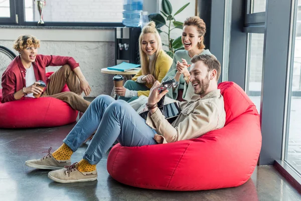 Group of friends sitting on bean bag chairs with coffee to go and playing guitar — Stock Photo