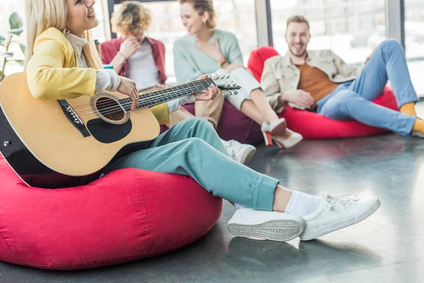 Selektiver Fokus einer fröhlichen Gruppe von Freunden, die auf Sitzsäcken sitzen und Gitarre spielen — Stockfoto