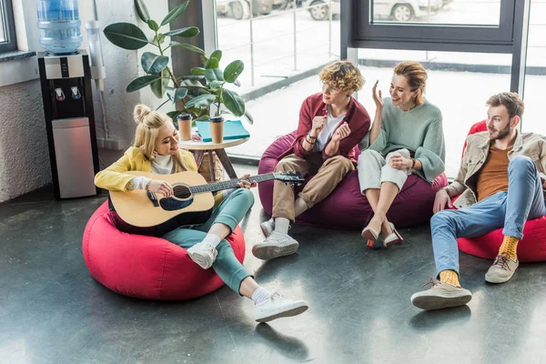 Grupo sonriente de amigos sentados en sillas bolsa de frijol y tocando la guitarra - foto de stock
