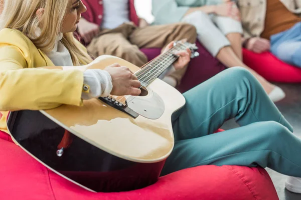 Обрезанный вид женщины, играющей на гитаре с друзьями, сидя на стуле из фасоли — стоковое фото