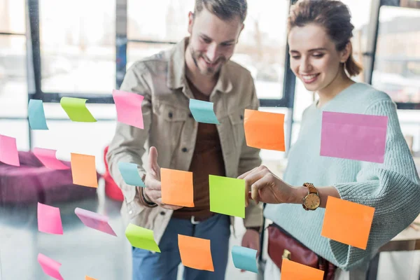 Sonriente hombre de negocios casual y mujer de negocios poniendo notas adhesivas de colores en la ventana de cristal en la oficina - foto de stock