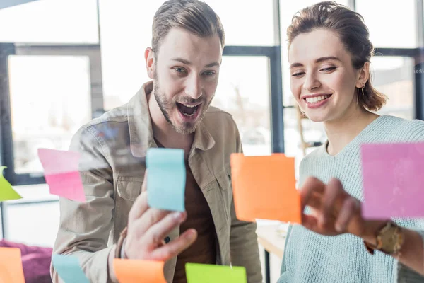 Sonriente hombre de negocios casual y mujer de negocios poniendo notas adhesivas de colores en la ventana de cristal en la oficina - foto de stock