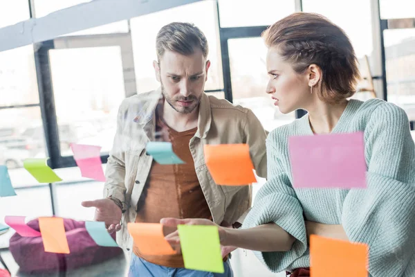 Confused casual businessman and businesswoman putting colorful sticky notes on glass window in office — Stock Photo