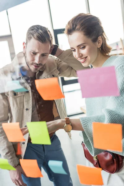 Casual businessman and businesswoman putting colorful sticky notes on glass window in office — Stock Photo