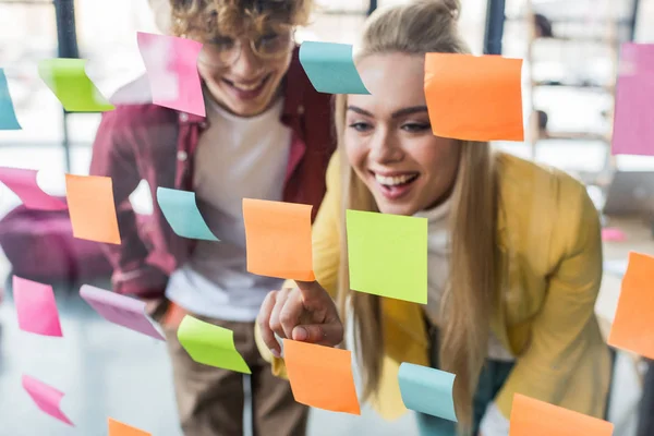 Happy casual businessman and businesswoman putting colorful sticky notes on glass window in office — Stock Photo