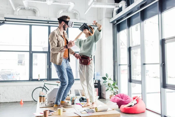 Diseñadores femeninos y masculinos que usan auriculares vr, haciendo gestos con las manos y teniendo experiencia de realidad virtual en la oficina loft - foto de stock