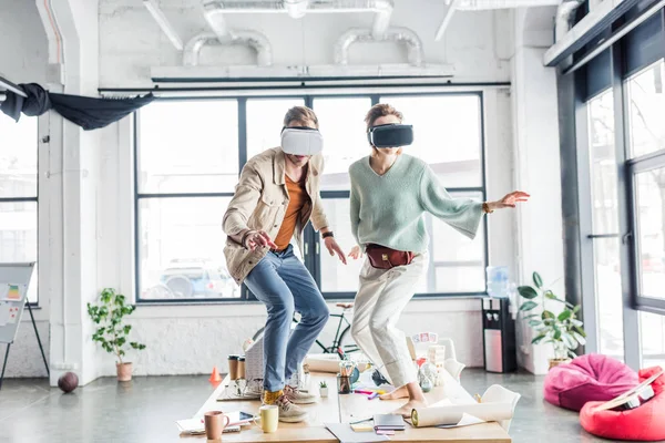 Arquitectos femeninos y masculinos que usan auriculares vr, haciendo gestos con las manos y teniendo experiencia de realidad virtual en la oficina loft - foto de stock