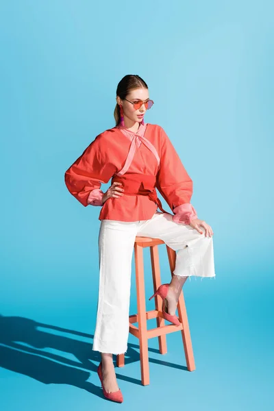 Femme séduisante posant dans des vêtements de corail vivant et des lunettes de soleil sur tabouret sur bleu — Photo de stock