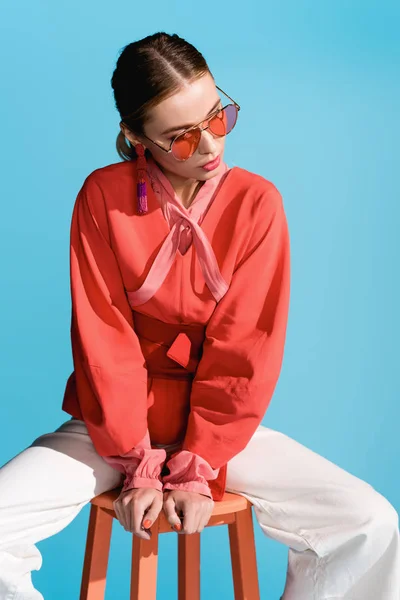 Femme élégante dans des vêtements de vie à la mode corail et lunettes de soleil posant sur tabouret isolé sur bleu — Photo de stock