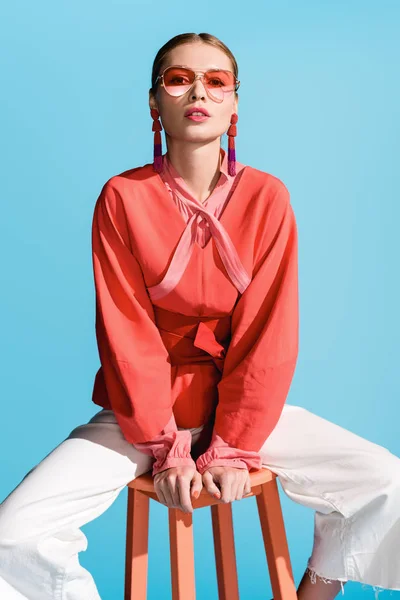 Модная модель в модной живой коралловой одежде и солнцезащитных очках, позирующих на стуле, изолированном от бирюзы — стоковое фото