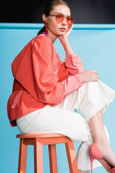 Модная девушка в модной живой коралловой одежде и солнцезащитных очках позирует на стуле на бирюзовой — стоковое фото