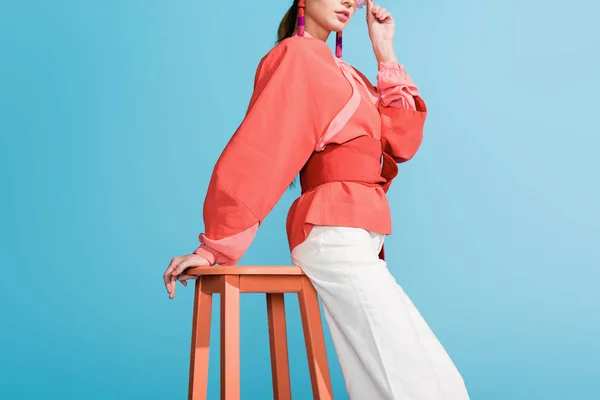 Vista recortada de la mujer en la ropa de coral de vida de moda posando en el taburete aislado en azul - foto de stock