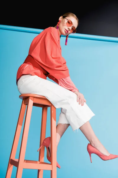 Belle femme dans des vêtements de corail vivant à la mode et des lunettes de soleil posant sur tabouret sur bleu — Photo de stock