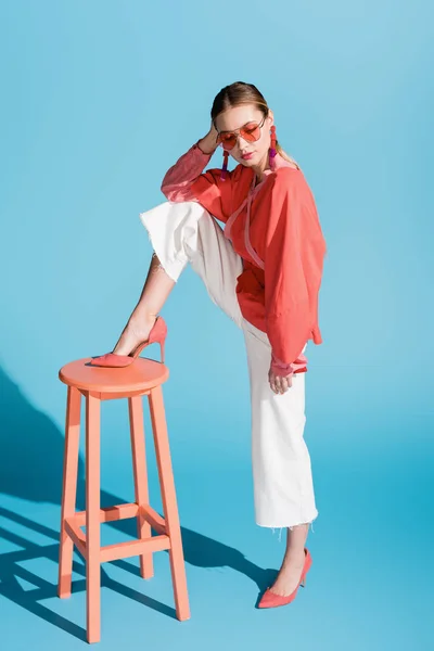 Modelo elegante em viver roupas de coral e óculos de sol posando em fezes em azul — Fotografia de Stock