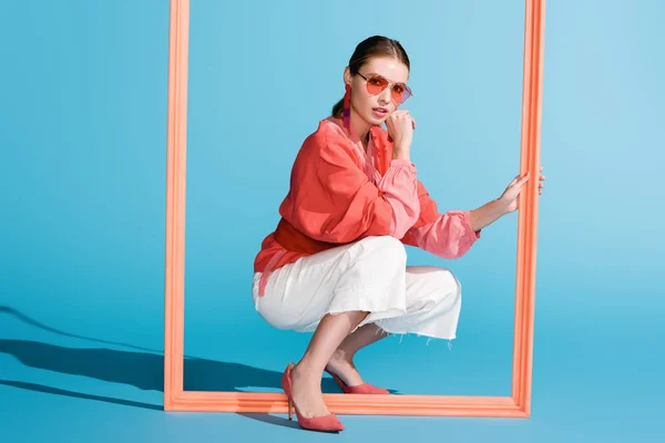 Modelo elegante em viver roupas de coral e óculos de sol posando com quadro grande em azul — Fotografia de Stock