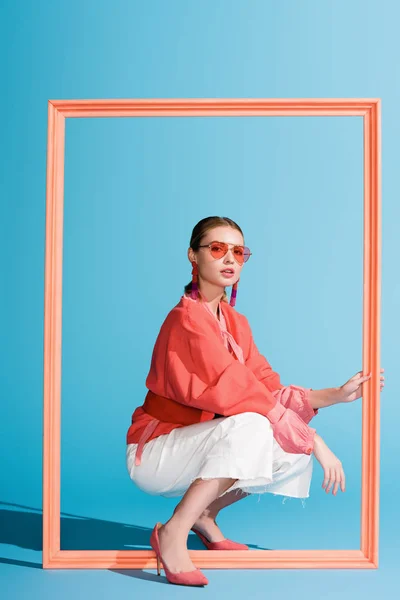 Модна дівчина в живому кораловому одязі і сонцезахисних окулярах позує з великою рамкою на синьому — стокове фото