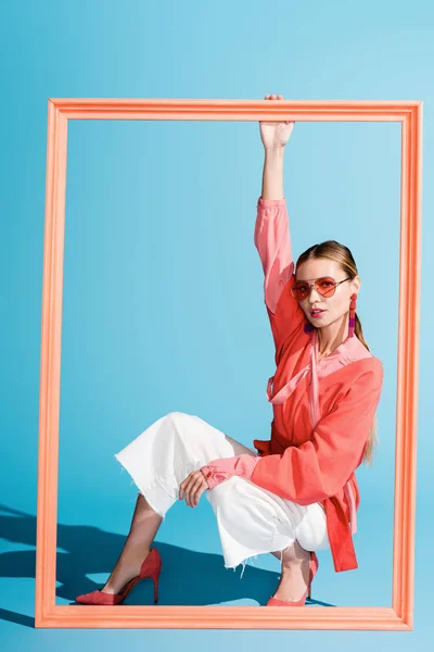 Beautiful young woman in living coral clothing and sunglasses posing with big frame on blue — Stock Photo