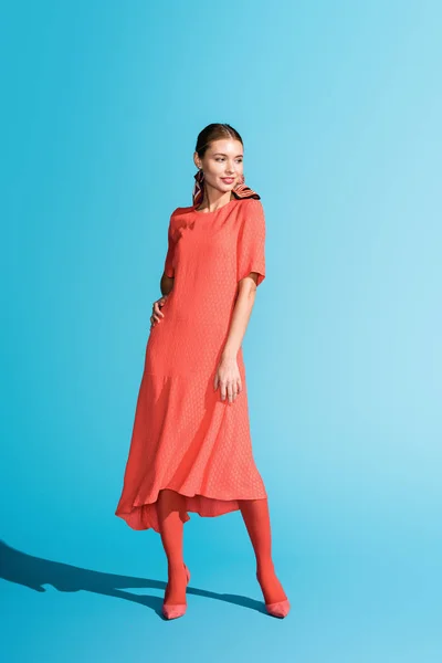 Модная фотосессия стильной улыбающейся модели в триумфальном живом коралловом платье, позирующей на голубом фоне — стоковое фото