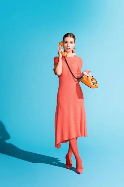 Приваблива модна модель в живій кораловій сукні, позує з поворотним телефоном на синьому — стокове фото