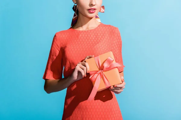 Vista recortada de la mujer de moda en vestido de coral vivo sosteniendo caja de regalo aislado en azul - foto de stock