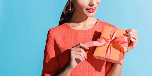 Vista recortada de la mujer de moda en vestido de coral vivo sosteniendo caja de regalo aislado en azul - foto de stock