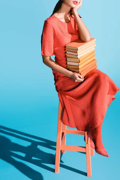Cropped view of stylish woman in living coral dress holding books and sitting on stool on blue — Stock Photo