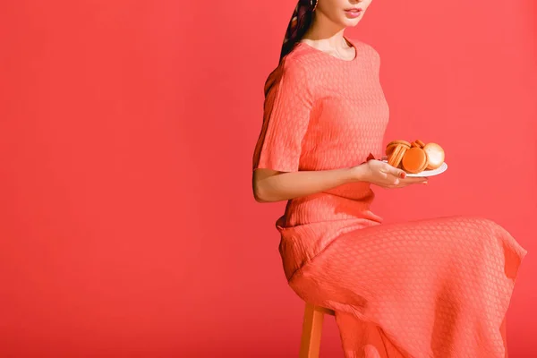 Vista cortada de menina elegante segurando placa com macaroons isolados em coral vivo. Pantone cor do ano 2019 conceito — Fotografia de Stock