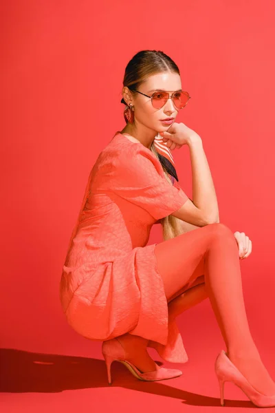 Élégant jeune femme posant dans la robe de corail vivant et des lunettes de soleil sur fond rouge — Photo de stock