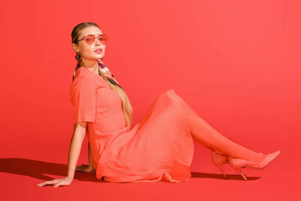 Modelo elegante posando em vestido de coral vivo e óculos de sol no fundo vermelho — Fotografia de Stock