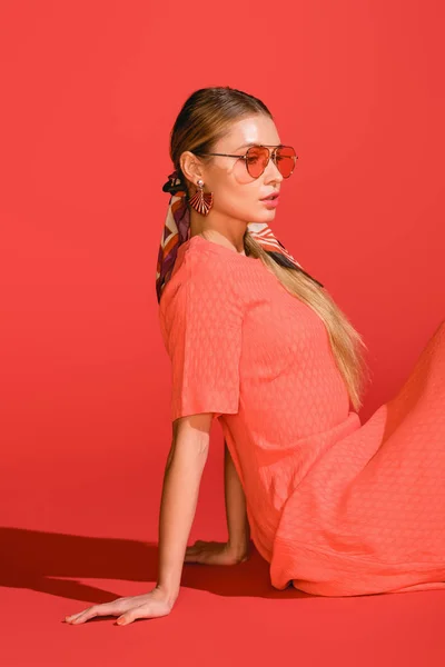 Modèle élégant à la mode posant en robe de corail vivant et lunettes de soleil sur fond rouge — Photo de stock