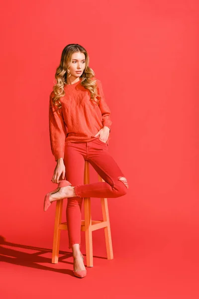 Élégante fille élégante posant dans des vêtements de corail vivant sur tabouret sur fond rouge — Photo de stock