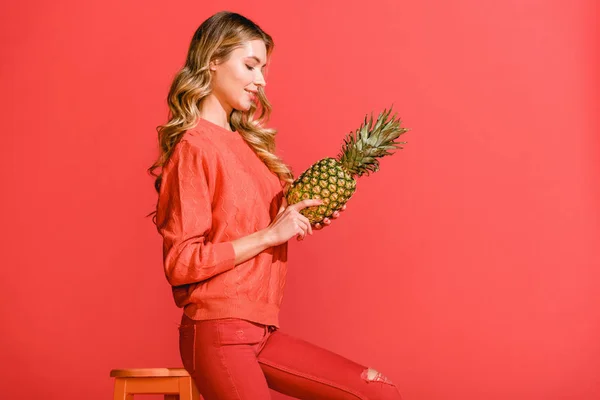 Attractive smiling girl holding fresh pineapple at stool isolated on living coral — Stock Photo
