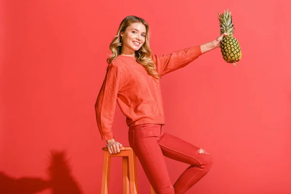 Stylish smiling woman holding fresh pineapple at stool isolated on living coral — Stock Photo